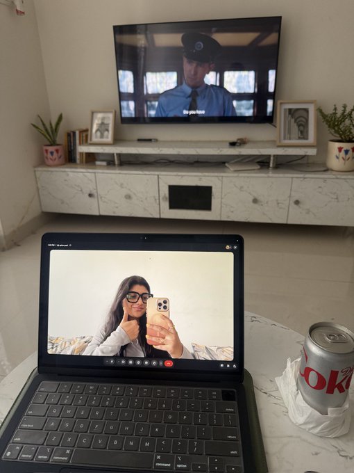 A tablet on a marble table shows a video call of a woman with glasses taking a selfie, while a TV in the background plays a show featuring a man in uniform and a Diet Coke can sits beside the tablet.
