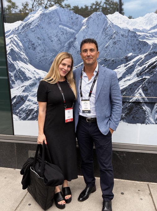 A man and woman pose together in front of a large mural of snowy mountain peaks; both wear lanyards with conference badges.