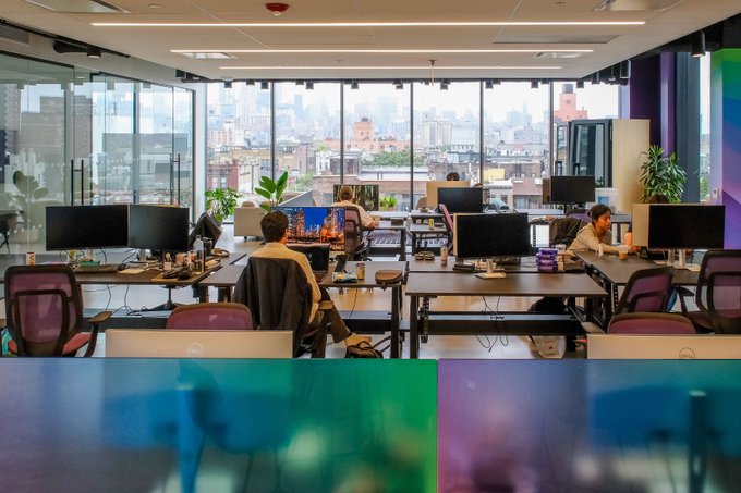 The image shows a large open office space filled with people working at desks. There are multiple individuals sitting at their respective workstations, using computers or other devices. The room has a modern design featuring glass walls that allow natural light to flood into the workspace. A few potted plants can be seen throughout the area, adding some greenery and life to the environment.

The post is about an upcoming event in which a group of people will be joining a new cohort on September 1st. The cohort consists of five individuals who will be working together for three months. They will have direct access to the ecosystem's top talent and meet with 90+ of NYC's top founders, investors, and operators.

The image is taken from a high angle, providing an overview of the office space and its occupants.