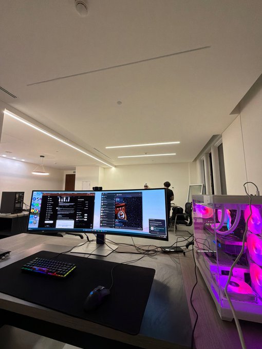 A dual-monitor trading setup displays market data and charts on a sleek desk, accompanied by an RGB mechanical keyboard, gaming mouse, and a transparent PC tower glowing with pink lighting in the background. The professional yet gaming-inspired workstation perfectly embodies the "Live from our trading floor" vibe, blending high-performance tech with an active trading environment.