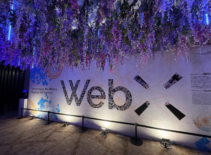 The image shows an exhibition wall with "Web" in large pixelated letters and the tagline "Connecting the Nodes, Beyond the Screen," adorned with cascading purple and blue wisteria flowers above, fitting the "Gmove WebX!" theme of a web-related event or experience.