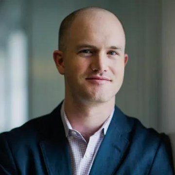 A professional headshot of a bald man wearing a dark navy blazer over a checkered shirt, smiling slightly at the camera against a blurred background.