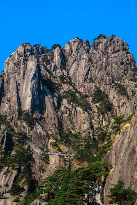 A photograph captures the rugged granite peaks of a mountain (resembling Huangshan) with pine trees clinging to the cliffs and a crowded stone staircase winding up the side under a clear blue sky.