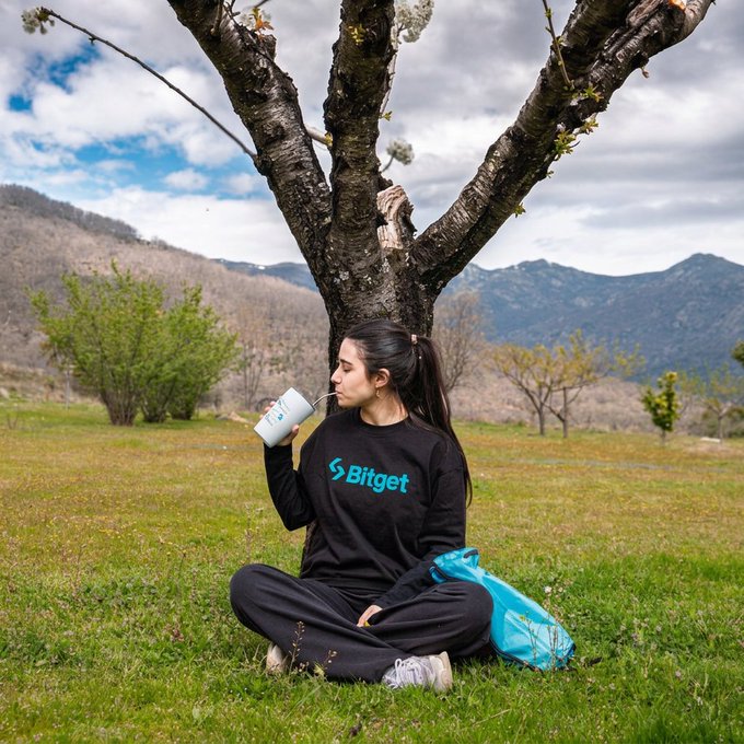 A young woman sits cross-legged under a blossoming tree in a grassy mountain meadow, peacefully sipping from a cup, with a "Bitget" sweatshirt visible and a bright blue bag beside her—suggesting a relaxed, well-earned weekend break. The serene outdoor scene embodies the carefree spirit of unwinding after a busy week.
