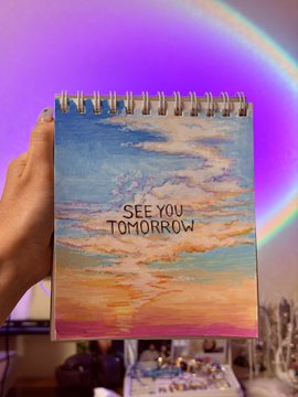 A hand holds a spiral notebook with a painted watercolor sky-sunset design and the text "SEE YOU TOMORROW", set against a vibrant purple and pink circular LED light background.