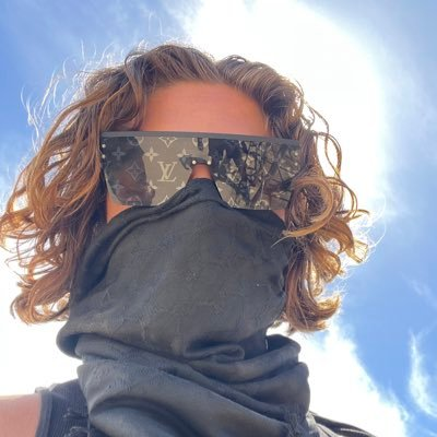 A low-angle selfie of a person with curly reddish-brown hair wearing designer sunglasses with a monogram pattern, a black face covering, and a dark scarf, set against a bright blue sky.