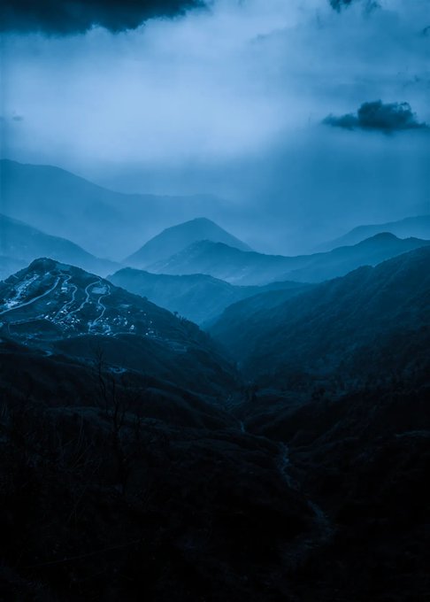 A misty, twilight mountain range with deep blue tones blankets the scene, where a small illuminated settlement nestles on a distant ridge beside winding roads, evoking the serene and remote landscape that complements the listed "Blue Mountains" NFT for sale at 0.065Ξ.
