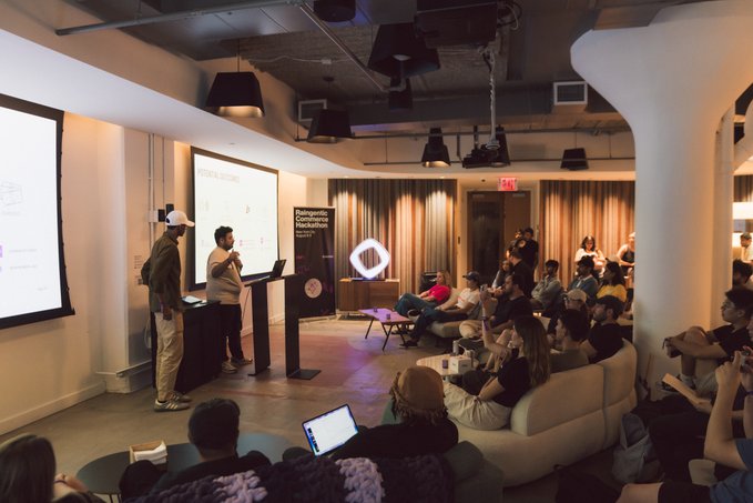 A presentation takes place in a modern lounge space with an audience seated on sofas watching speakers at a podium with 'Poimorphic Crypto Hackathon' signage.