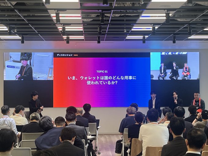 A panel discussion titled 'ディスカッション TOPIC' featuring Japanese text 'いま、ウォレットは誰のどんな仕事に使われているか?' (Who uses wallets and for what purposes?) is shown on a large screen, with panelists seated before an audience.