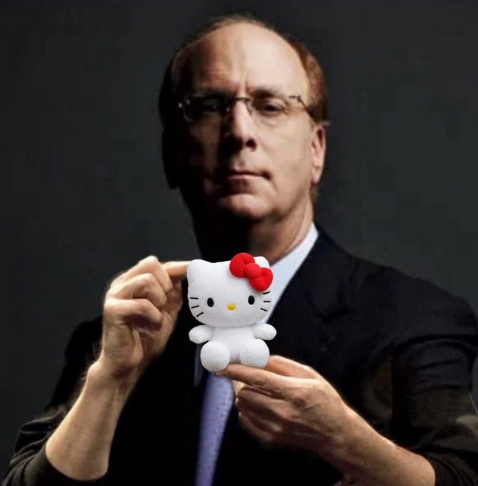 A man in a dark suit and glasses holds up a Hello Kitty plush figure against a dark background.