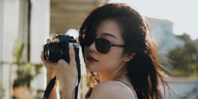 A woman with long hair wearing sunglasses holding a camera, possibly taking a picture of herself or someone else. The photo has been edited to make her appear as if she's just woken up from sleep.