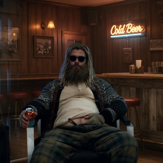 A long-haired bearded man wearing sunglasses and a patterned cardigan sits in a chair holding a canned drink, with a neon 'Cold Beer' sign visible in the wood-paneled bar behind him.