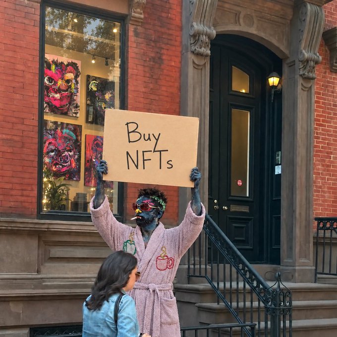 A person with colorful face paint, wearing a pink robe and red sunglasses, holds up a handwritten cardboard sign reading "Buy NFTs" on a city sidewalk, with artwork displayed in a window behind them.