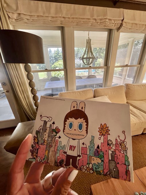 A hand holding a card with colorful illustrations of a cartoon character and flowers, photographed in a living room setting.