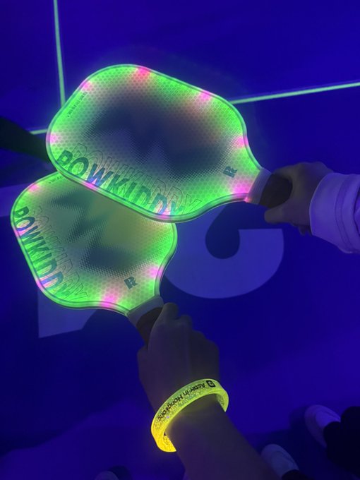 Two hands wearing yellow "Boombox" wristbands hold up light-up pickleball paddles glowing green and pink under blacklight, displaying the "BOOMBOX" brand name on their translucent faces.