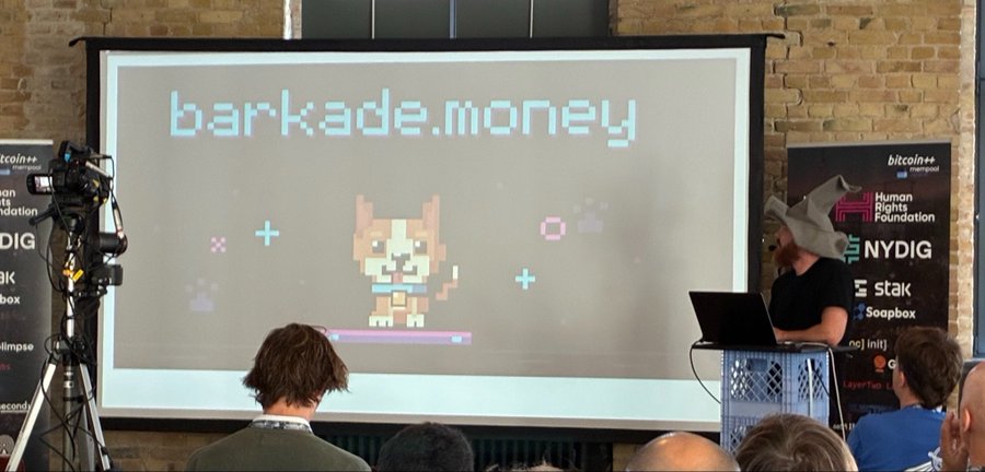 A conference presentation showing a slide titled 'barkade.money' with a pixel art dog character, with a speaker wearing a similar paper hat visible on the right and audience members in the foreground.