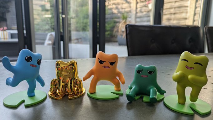 Photo of five small quirky figurines arranged on a table: a blue dancing character, a gold metallic figure, an orange character, a green blob-like character, and a yellow smiling character, each on a green base.