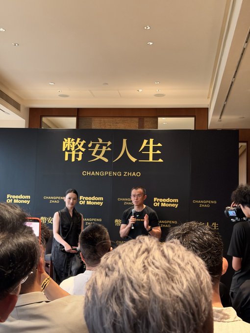 Changpeng Zhao (CZ) speaks at a book event for 'Freedom of Money' (幣安人生), wearing a black t-shirt, with a woman standing beside him and a crowd of attendees watching.