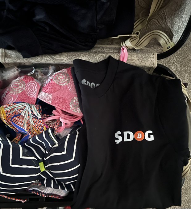 An open suitcase filled with packed clothes, prominently featuring a black t-shirt with a '$DOBG' logo where the 'O' is a Bitcoin symbol.
