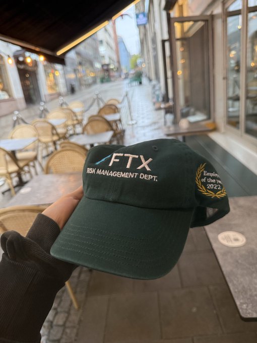 A hand holds up a dark green 'FTX RISK MANAGEMENT DEPT. Employee of the year 2022' embroidered hat in front of a cafe patio.