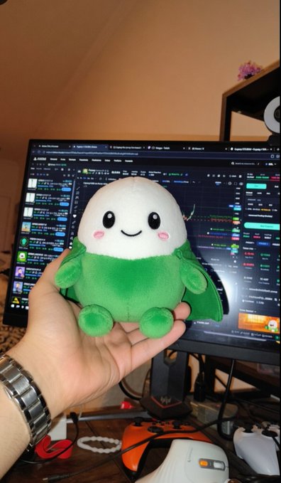 A hand holding a small green and white plush toy in front of a computer monitor displaying a cryptocurrency trading platform. A watch is visible on the wrist.