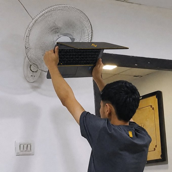 A man reaches up to place or adjust a black laptop near the blades of a white wall-mounted electric fan, with a decorative yellow and black panel on the wall nearby.