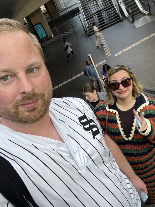 The image features a man wearing a baseball jersey with "S" on his shirt, standing next to a woman who is also dressed up for a special occasion. They are both posing together for a picture at an indoor location, possibly a mall or a public building. The background of the photo shows stairs and escalators, indicating that they might be in a shopping center or a similar environment. The image includes text from a post on social media, which reads: "GM Have an amazing sunday Oh and to have an amazing sunday you ofcourse use Solflare".