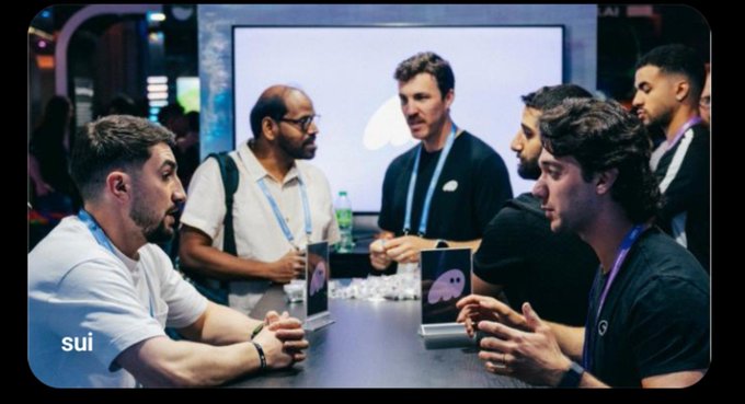A group of men sit around a table in discussion, with the "sui" logo visible in the bottom left corner and a Discord logo on a small display, suggesting a tech or blockchain-related conference meeting.