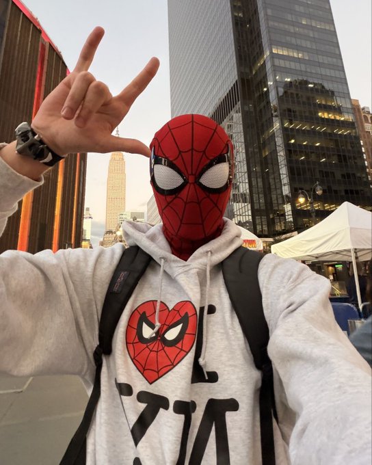 A person wearing a Spider-Man mask and a gray hoodie with a Spider-Man heart design makes a hand gesture in front of tall city buildings.