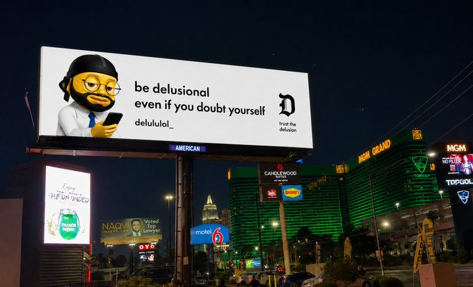 A nighttime photograph of a billboard in Las Vegas featuring a 3D monkey character and the motivational text 'be delusional even if you doubt yourself delulu.lol', with the MGM Grand in the background.