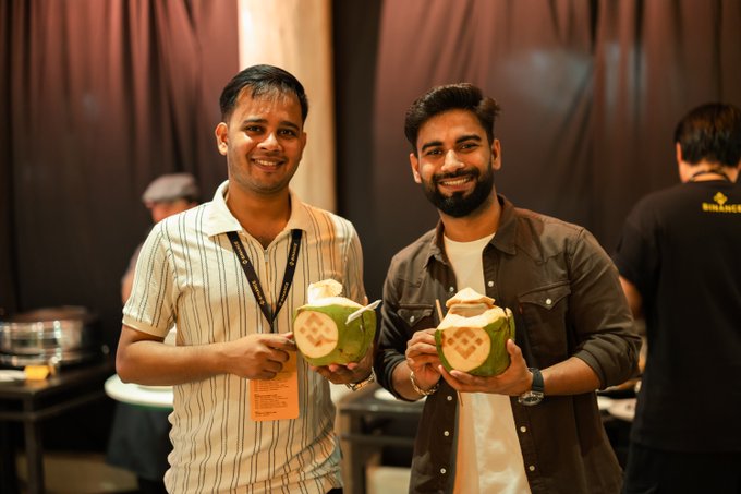 Two smiling men pose together at an event, each holding a fresh green coconut with a straw, with one wearing a lanyard with an orange name badge.