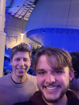 Two men take a selfie in front of a large illuminated blue dome-like structure at night.