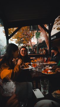 A group of people enjoy drinks and a meal at a wooden picnic table under a covered outdoor structure during sunset or early evening.