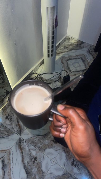 A hand holds a dark mug filled with a light brown beverage, likely coffee or tea with milk, next to a tall tower fan on a marble-patterned floor with visible cables.