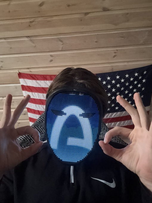 A person wearing a black hoodie with a Nike logo, holding up both hands in "OK" gestures, with their face obscured by a blue LED mask displaying a face graphic; an American flag hangs on the wooden wall behind them.