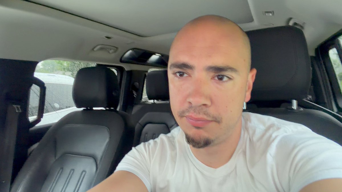 The image shows a bald man wearing a white t-shirt, sitting inside a car. The man appears to be looking at the camera while driving down the road. There are several other cars visible outside of the frame, indicating that they might be in an urban setting or on a busy street. The post about Bitcoin and XRPL speculations is related to the image as it suggests that the growth of Bitcoin could potentially impact the automotive industry due to the use of stablecoins for transactions within the car manufacturing supply chain.