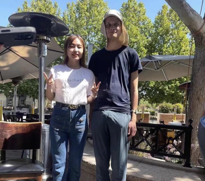 A casual photo of a young woman in a white shirt and jeans making peace signs standing next to a very tall young man in a black shirt and cap, posed outdoors at a patio area.