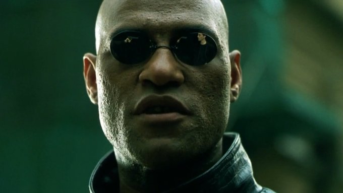 A close-up portrait of a bald man wearing dark oval sunglasses with a serious expression.