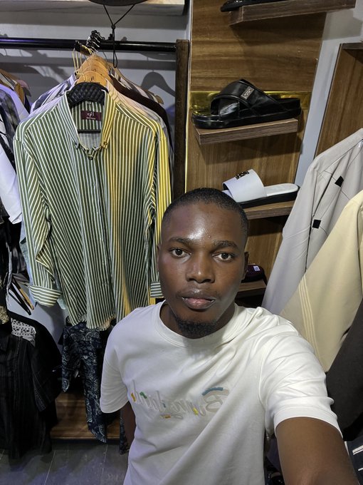 A man in a white t-shirt taking a selfie inside a clothing store, with a striped shirt hanging on a rack and sandals displayed on wooden shelves behind him.
