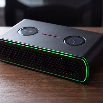 A black electronic device with green LED lighting around its edges, featuring two circular dials on top and a perforated grill, sitting on a wooden surface.