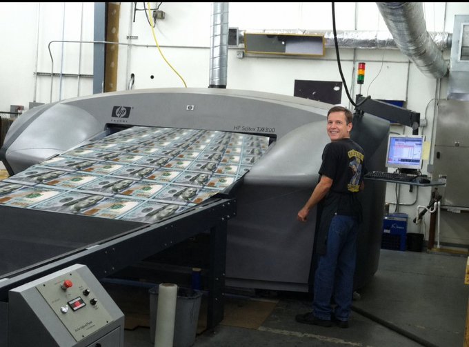 The image features a man standing next to a large machine that prints money, likely an HP printer. He appears to be smiling as he looks at the machine. The text "Crypto is backed by nothing" can also be seen in the image. In addition, there are two other people present in the scene, one of whom has a computer monitor and keyboard nearby.