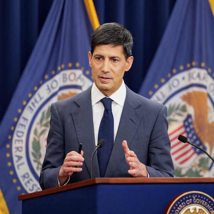 A man in a blue suit and tie speaks at a podium bearing the Federal Reserve Board of Governors seal, with Federal Reserve flags behind him.