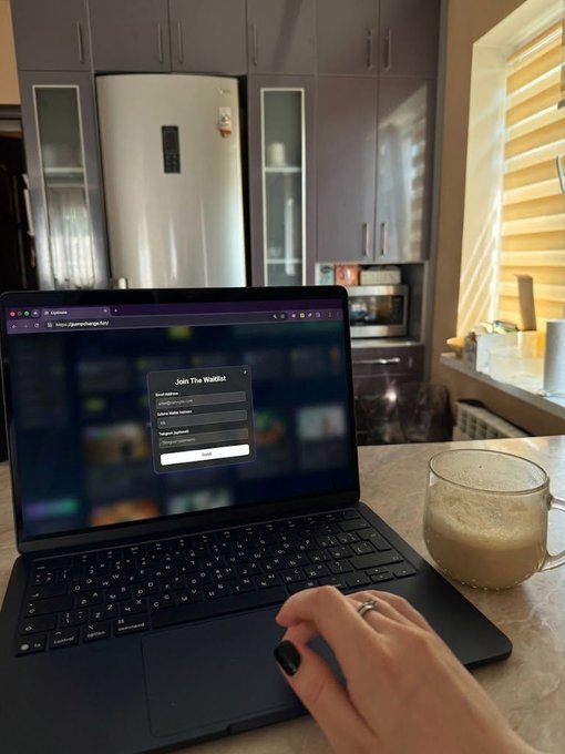 A person with dark nail polish and a ring uses a laptop on a kitchen counter showing a 'Join The Waitlist' signup page, with a frothy drink beside it and kitchen cabinets and a refrigerator in the background.