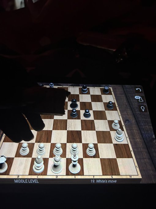 A photo of a hand playing chess on a tablet screen showing 'MIDDLE LEVEL 19. White's move' with wooden chess pieces rendered digitally.