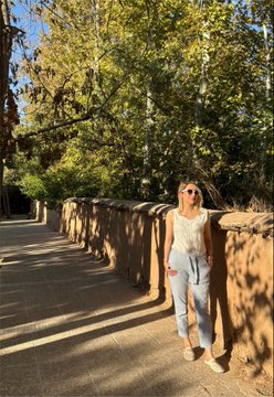 A woman in jeans and a white top leans against a sunlit stone wall on a tree-lined path, embodying the bright "between sun and rain" Friday energy referenced in the caption. The relaxed, golden-hour vibe complements the cheerful "$XTZ" crypto ticker and casual Good Morning post.
