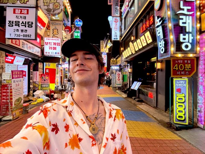 A young man in a black cap and floral shirt smiles with eyes closed in a selfie taken on a vibrant Korean street filled with neon signs advertising restaurants, coffee shops ("THEBOX," "THE HOLIC"), and karaoke venues ("노래방"), with one sign prominently displaying "40분" and another showing "1000." The post's theme of "clarity" connects to his serene, eyes-closed expression—capturing a moment of peaceful awareness amid the visually overwhelming, sign-saturated alley.