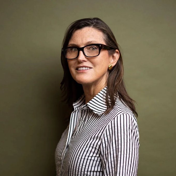 A portrait of a smiling woman with long brown hair wearing black-rimmed glasses and a black-and-white striped collared shirt, photographed against an olive green background.