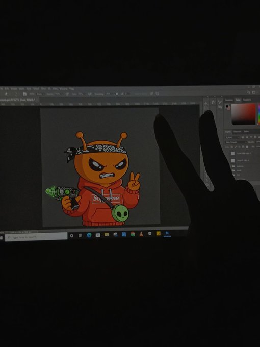 A computer screen displaying digital art of an orange alien-like character in a red Supreme hoodie holding a gun, with a hand making a peace sign in the foreground.