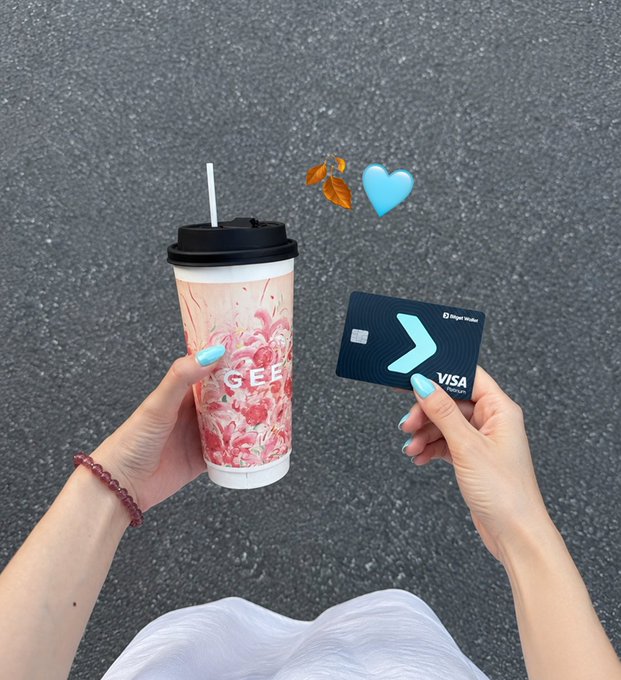 A hand with pastel blue nails holds a pink floral "GEE" branded milk tea cup alongside a black "FigerWallet VISA" debit card, set against an asphalt background and accented with a blue heart and autumn leaf emoji. The image perfectly captures the post's theme of enjoying a seasonal milk tea treat paid for using a digital asset-backed Visa card, blending cozy autumn vibes with modern fintech convenience.