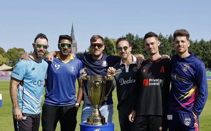 Six men pose with a large golden trophy on a cricket field, wearing various team jerseys including Glasgow Cosmic, Docker, Private Invest, and Hygrade, suggesting a tournament celebration photo. The image relates to the post's mention of the ETPL cricket league and the user traveling to Amsterdam to support the Glasgow Cosmic team, though the caption notes the image appears to show a different event as the men's team jerseys don't directly match the user's cricket context.